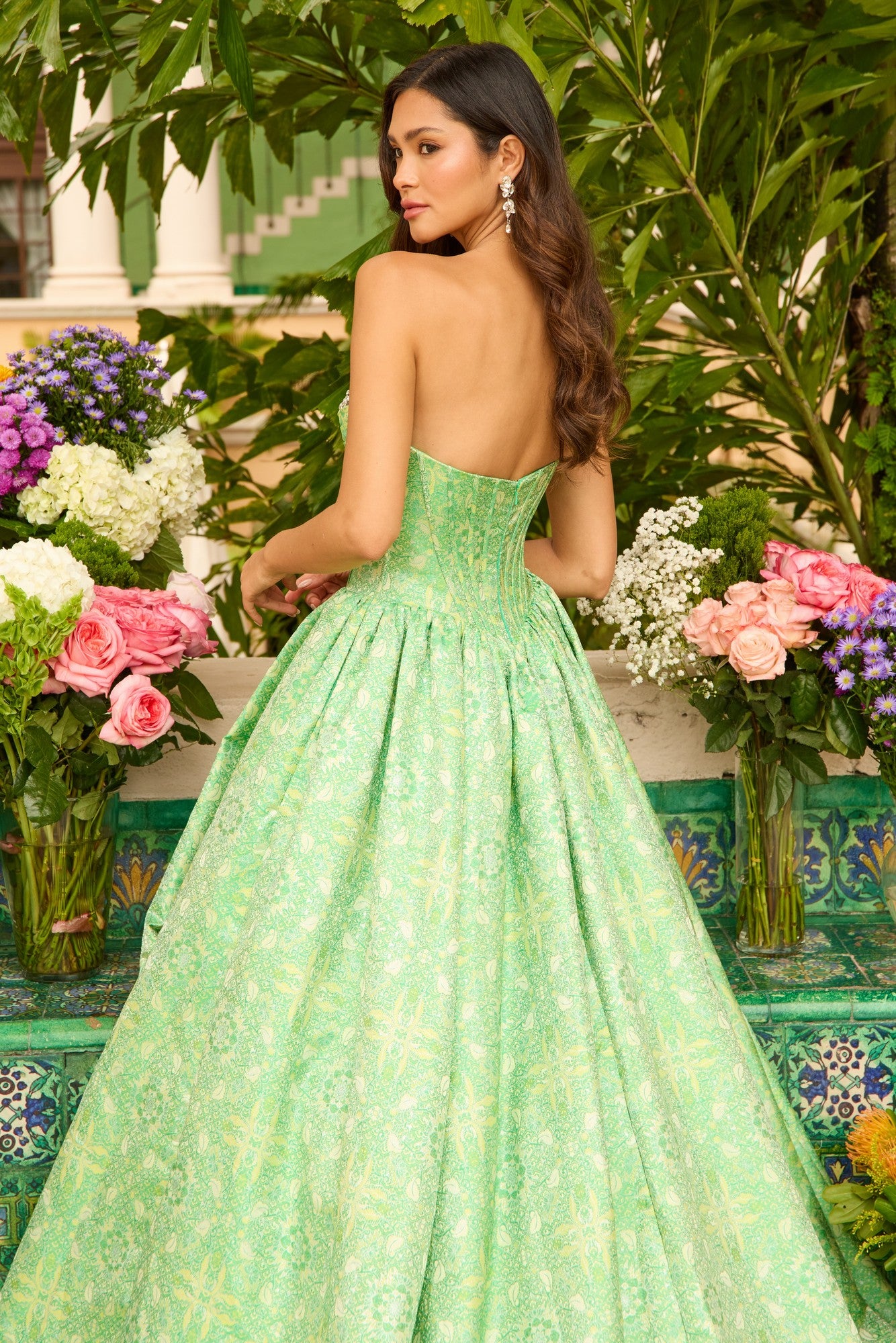 A woman in the Ava Presley 42318 long prom dress, featuring a strapless corset bodice, stands outdoors among colorful flowers with her back to the camera.